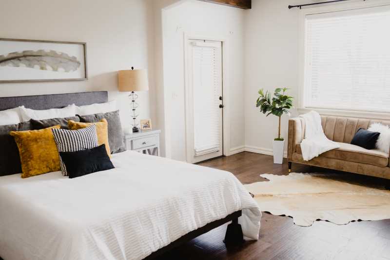 a white bed with cushions, a painting, a lamp, a plant, and a sofa