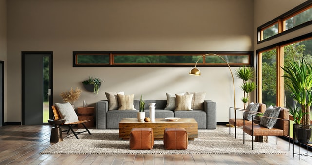 A big living room with large open windows, a grey couch surrounded by plants, and a brown coffee table